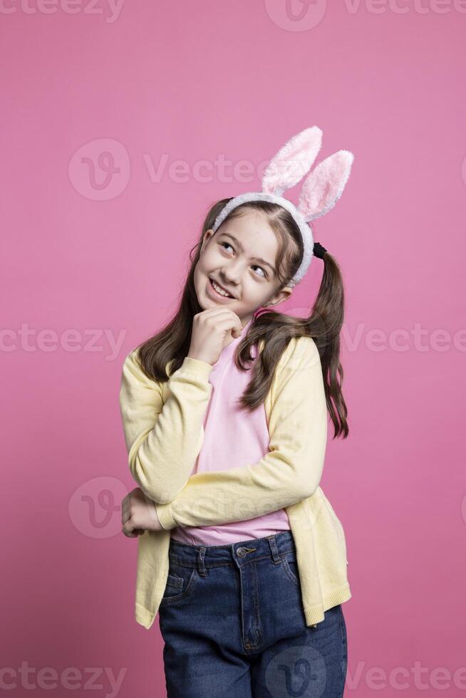 Little toddler smiling in studio thinking about presents, being excited about easter holiday celebration. Joyful schoolgirl with bunny ears and pigtails being pensive on camera, sweet kid. photo