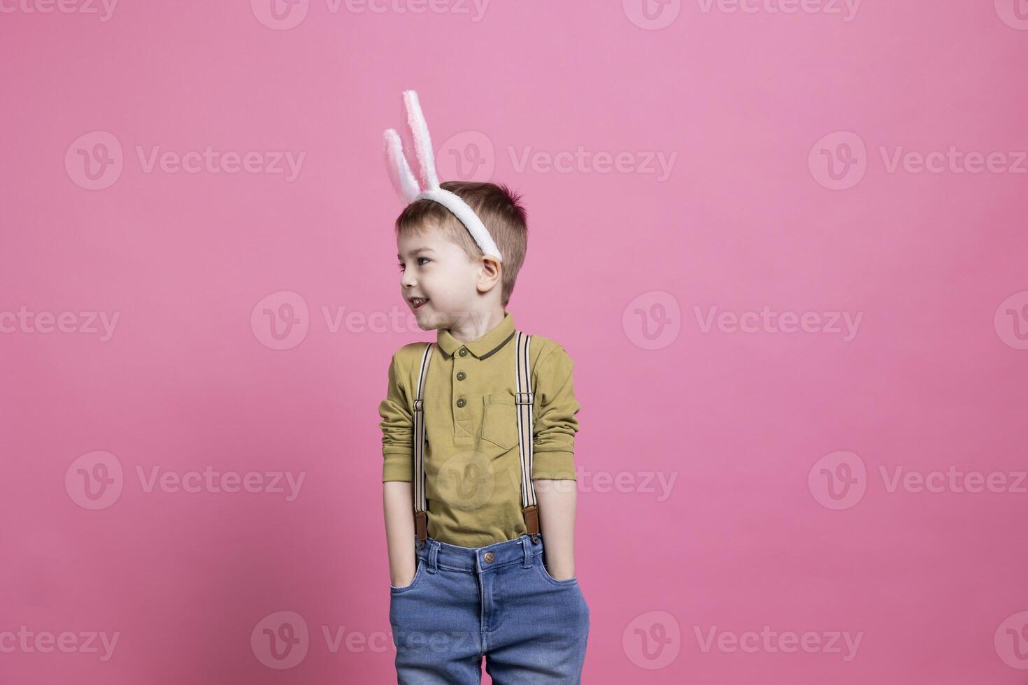 Cheerful young kid smiling and wearing fluffy bunny ears on camera, feeling joyful about easter festivity celebration and standing against pink background. Adorable small boy with colorful outfit. photo