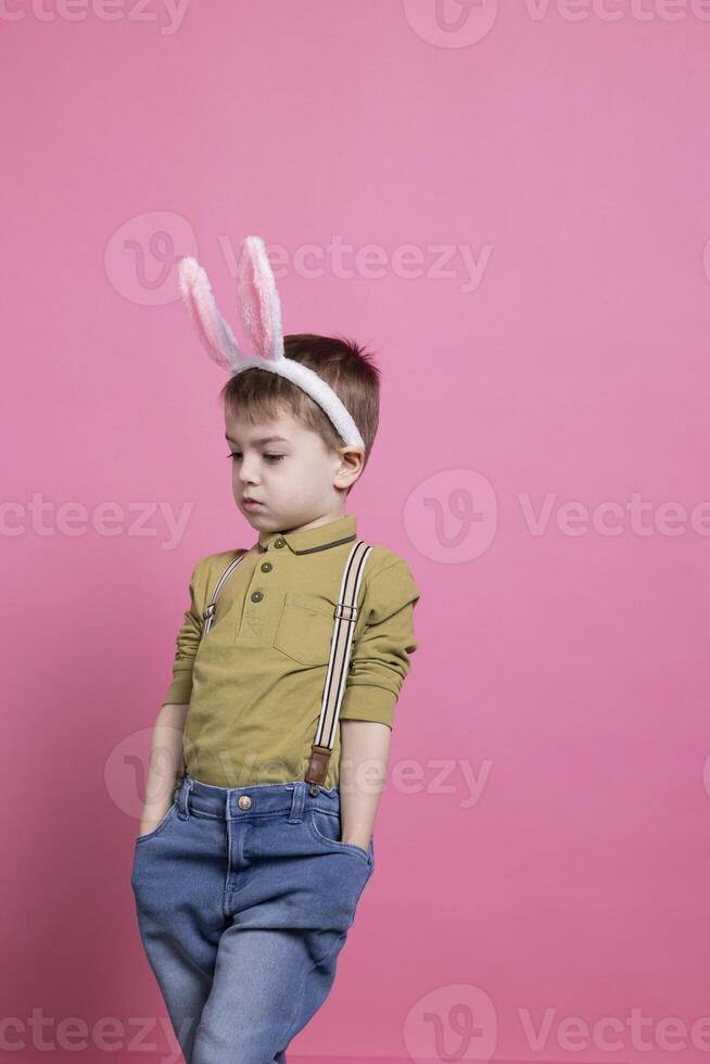 Lovely small boy wearing fluffy bunny ears and posing with confidence on camera, feeling excited about easter celebration and standing against pink backdrop. Cheerful preschooler with adorable outfit. photo