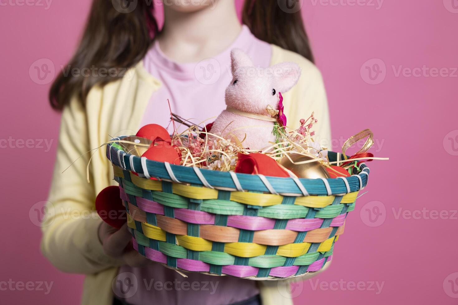 Small child presenting easter decorations and toys in a basket, holding colorful spring festive arrangements in front of camera. Young positive girl smiling in studio with adorable decor. Close up. photo