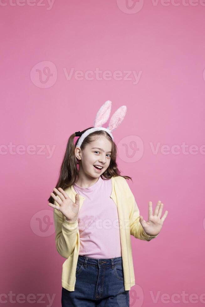 Little child acting excited and cheerful about easter festivity, posing with confidence against pink background. Young small kid wearing bunny ears and pigtails, feeling cheerful and carefree. photo