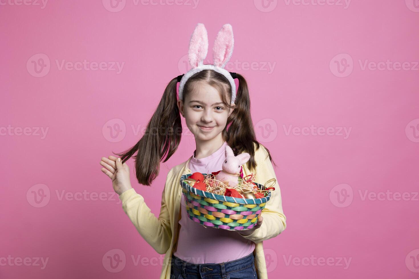 Little child with bunny ears holds Easter toys and decorations, evoking the enchantment of the season. Cute girl with adorable smile showing a rabbit and eggs in a basket on camera. photo