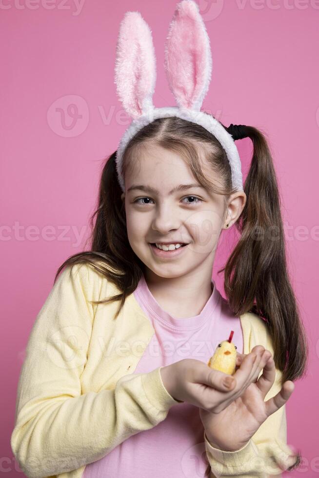 Small joyful girl presenting a fluffy chick toy in pink studio, adorable young child with golden stuffed toy over colored background. Little kid with festive bunny ears for seasonal celebration. photo