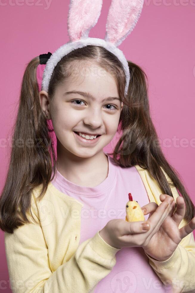 Young cute kid holding a stuffed chick in front of camera, positive excited girl feeling happy about easter celebrations. Small child with fluffy bunny ears smiling over pink background. photo