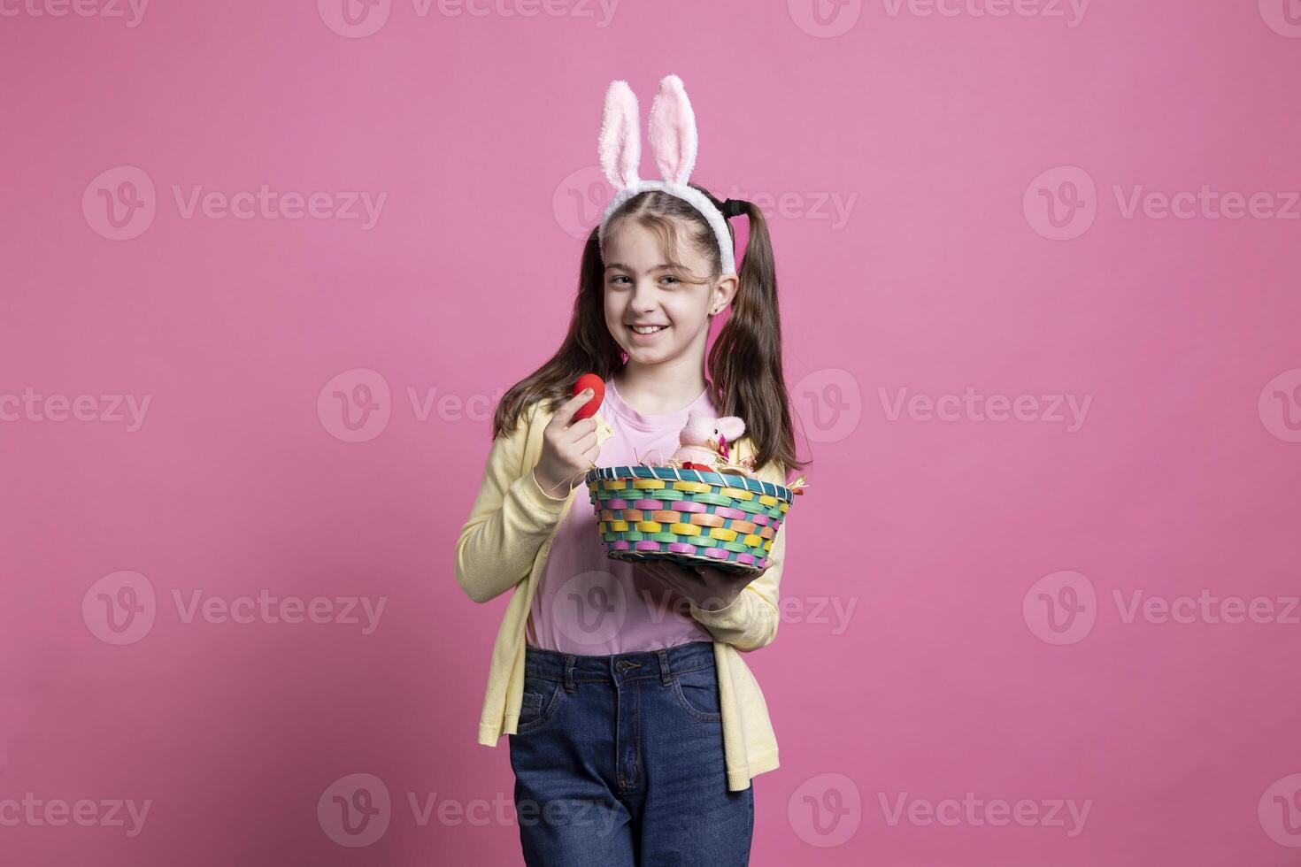 adorable alegre niño participación vistoso huevos y un relleno Conejo en un Pascua de Resurrección acuerdo, posando en rosado estudio. joven sonriente niña con conejito orejas participación un cesta con encantador primavera adornos foto