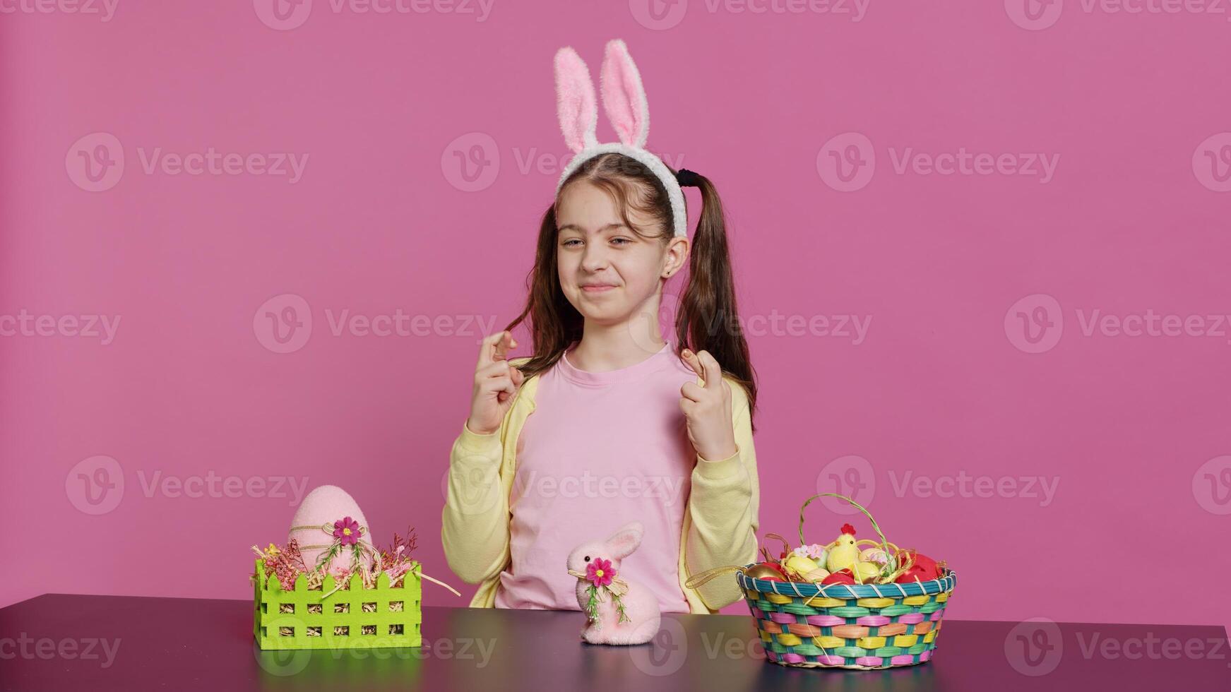 Small cheerful kid posing with fingers crossed in studio, sitting at a table filled with easter festive ornaments. Young schoolgirl hoping for luck and fortune during spring holiday celebration. Camera A. photo