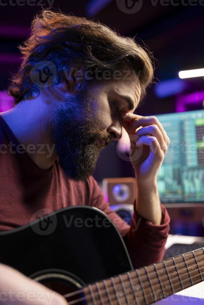 Stressed sleepy artist practicing acoustic instrument in his home studio, recording new tunes and editing it in post production. Young artist playing guitar with frustration, deals with burnout. photo