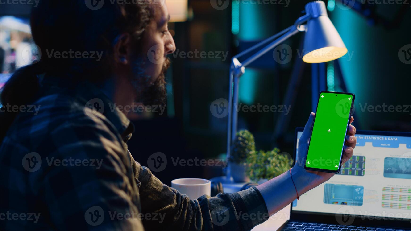 Self employed man in home office checks statistics on laptop, crosschecking with data on chroma key smartphone. Remote worker holding isolated screen phone, analyzing business graphics, panning shot photo