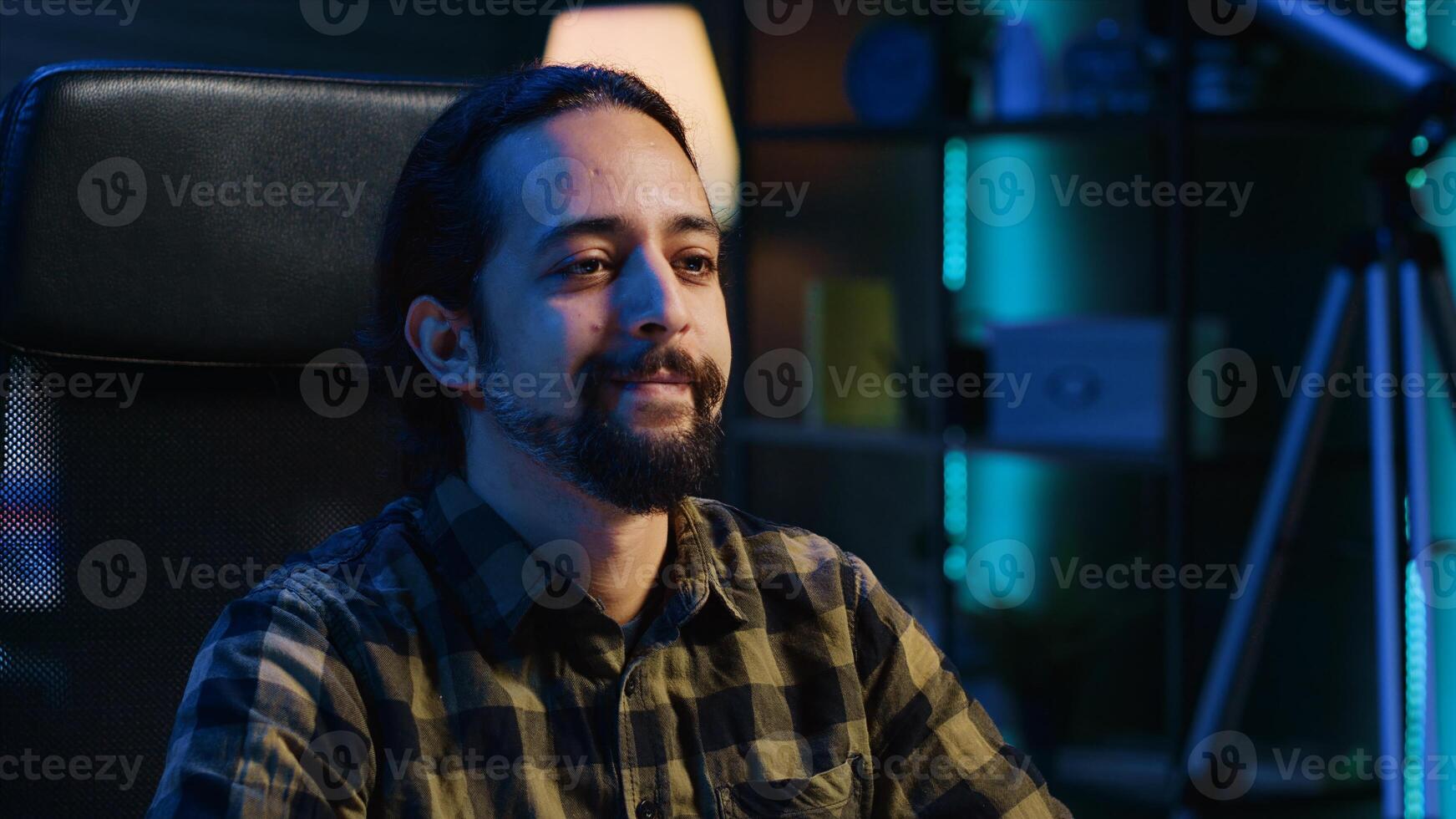 Smiling caucasian man sitting on chair at home office desk in neon illuminated apartment late at night. Cheerful person in RGB lit living room enjoying leisure time, camera A close up shot photo