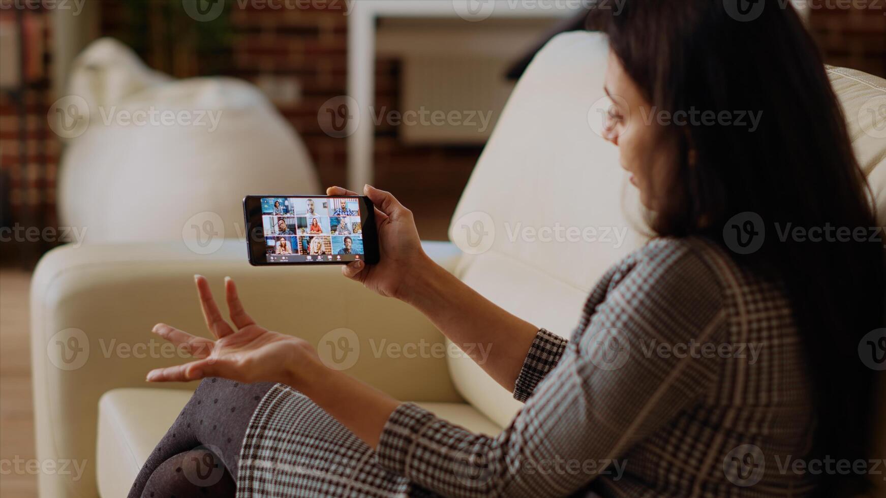 Management executive using smartphone to delegate project tasks to employees during videoconference meeting. Manager at home holding internet videocall on phone with diverse team photo