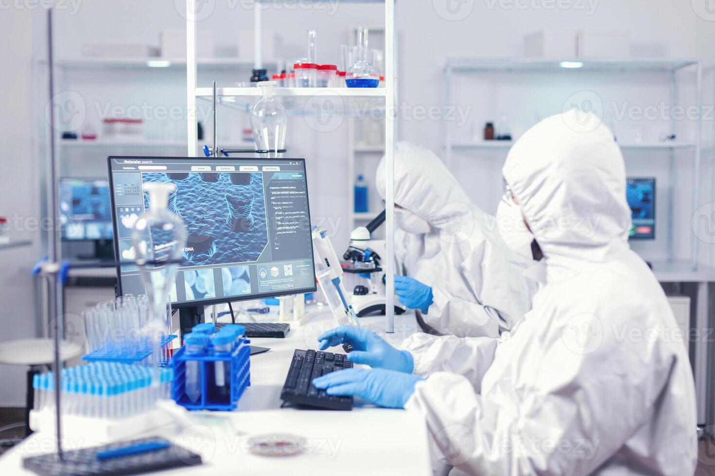 químico en ppe traje mecanografía en teclado comprobación virus desarrollo en equipado laboratorio. médico ingeniero utilizando computadora durante global pandemia con coronavirus vestido en mono. foto