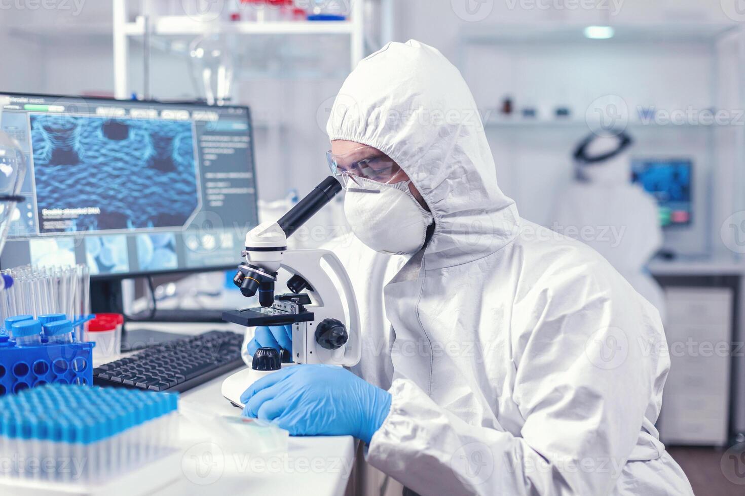 Doctor researching and evaluating virus using microscope in laboratory wearing ppe. Scientist in protective suit sitting at workplace using modern medical technology during global epidemic. photo