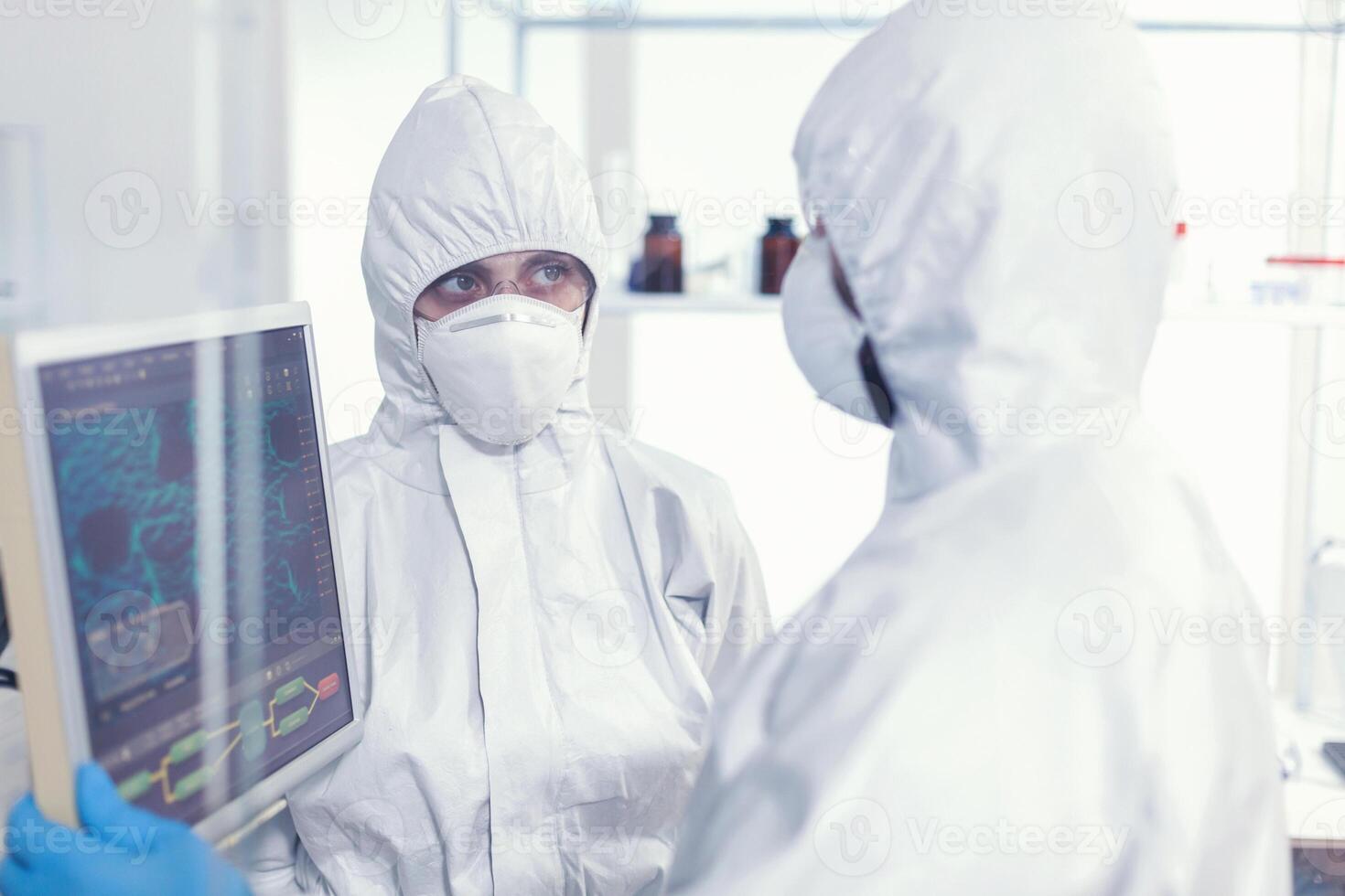 científicos compañeros de trabajo en bata que se discute acerca de virus evolución en equipado laboratorio señalando en computadora escritorio. doctores examinando virus evolución utilizando alto tecnología investigando diagnóstico en contra covid19 foto