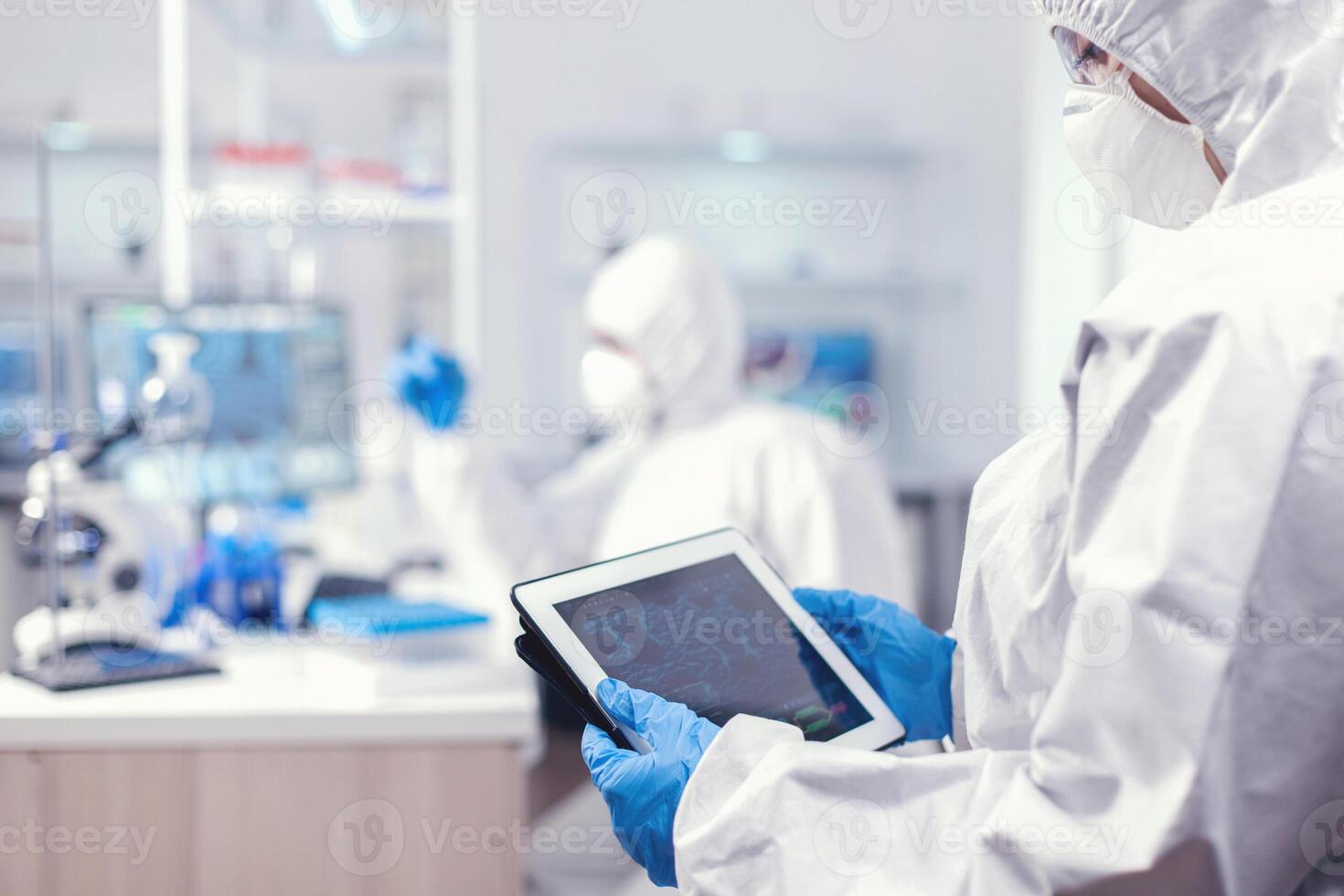 Back view of doctor in protection suit analyzing virus evolution looking on digital tablet. Team of scientists conducting vaccine development using high tech technology for researching treatment against covid19 photo