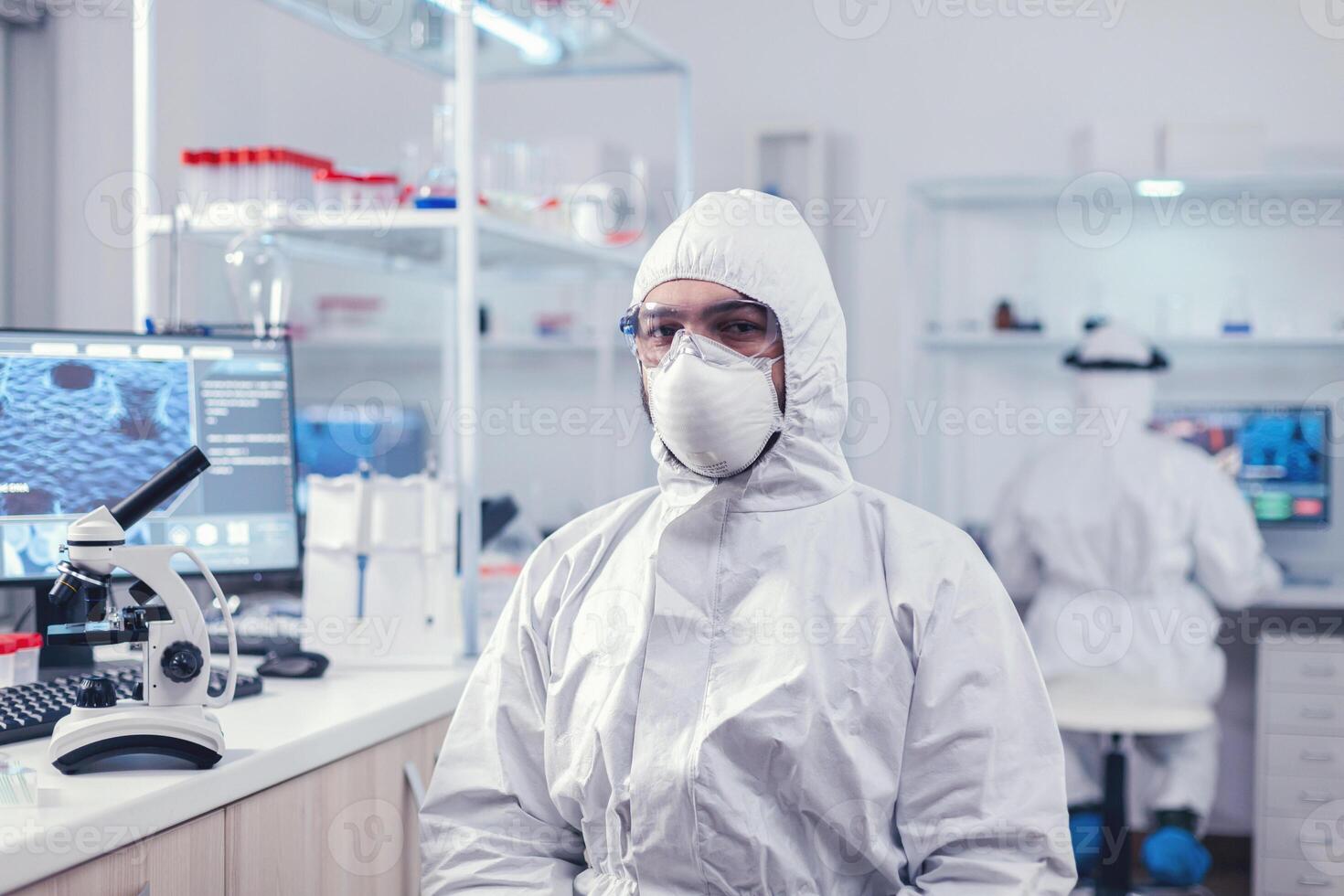 Chemist feeling tired from work for a long time in laboratory sitting at workplace. Overworked researcher dressed in protective suit against invection with coronavirus during global epidemic. photo