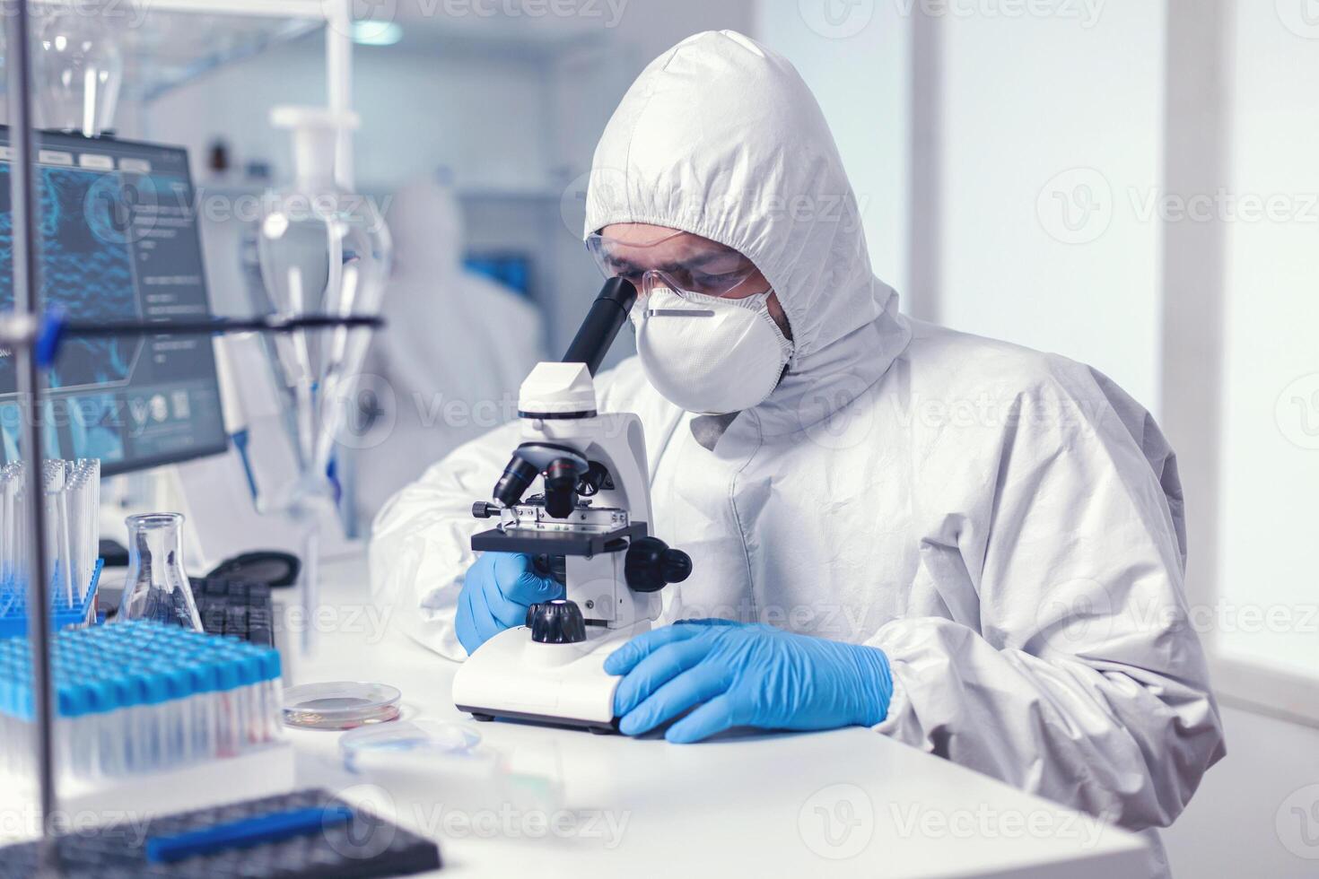 Microbiologist with protection glasses using microscope dressed ppe suit. Virolog in coverall during coronavirus outbreak conducting healthcare scientific analysis. photo