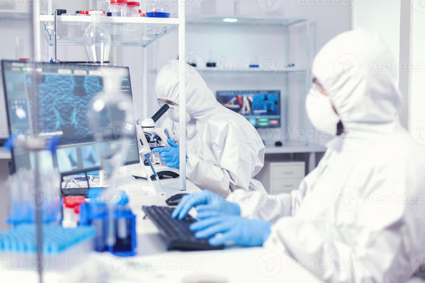 Woman researcher carrying out scientific covid-19 test using microscope. Medical engineer using computer during global pandemic with coronavirus dressed in coverall. photo