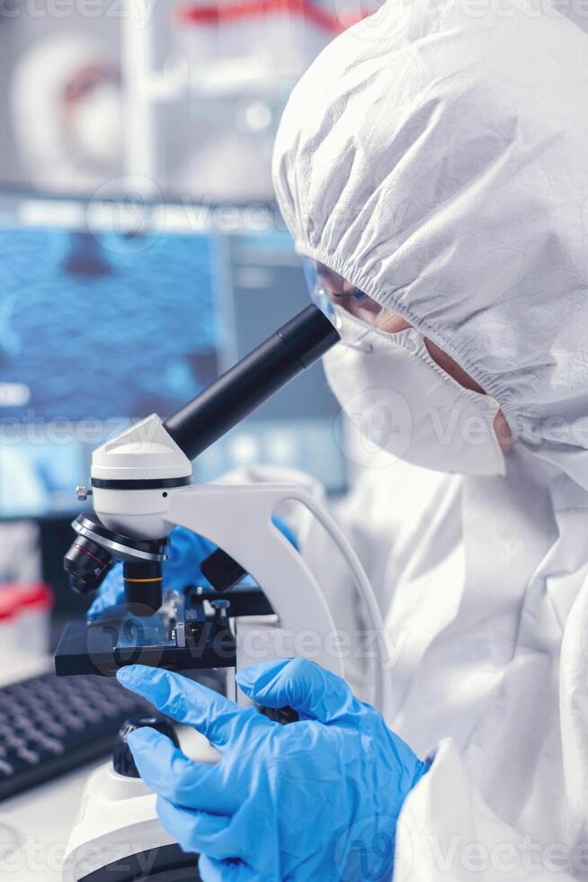 Medical scientist analysing sample on glass slide looking through microscope. Chemist researcher during global pandemic with covid-19 checking sample in biochemistry lab photo