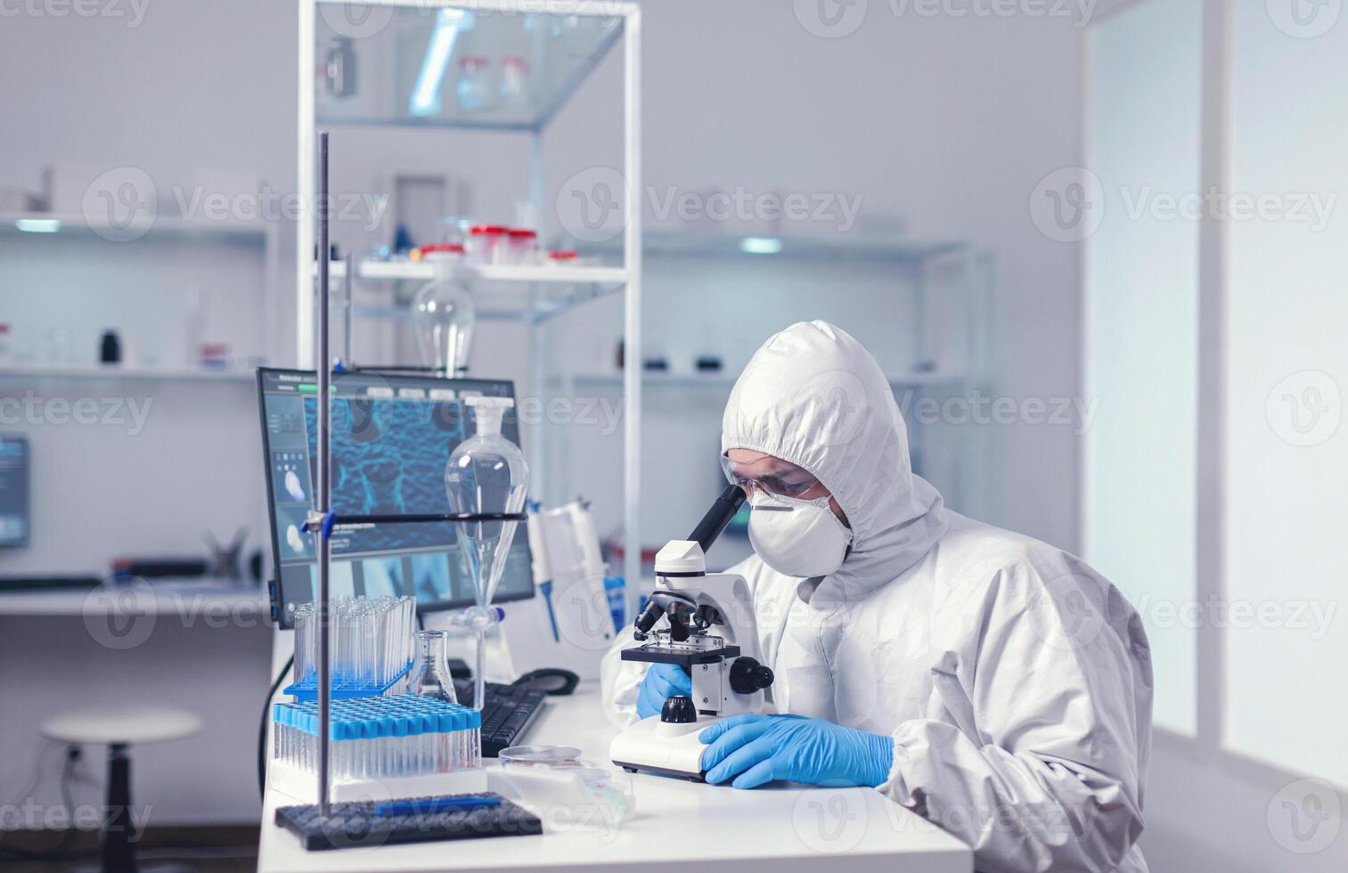 Scientist studying new virus in laborator using microscope dressed in protection suit. Virolog in coverall during coronavirus outbreak conducting healthcare scientific analysis. photo