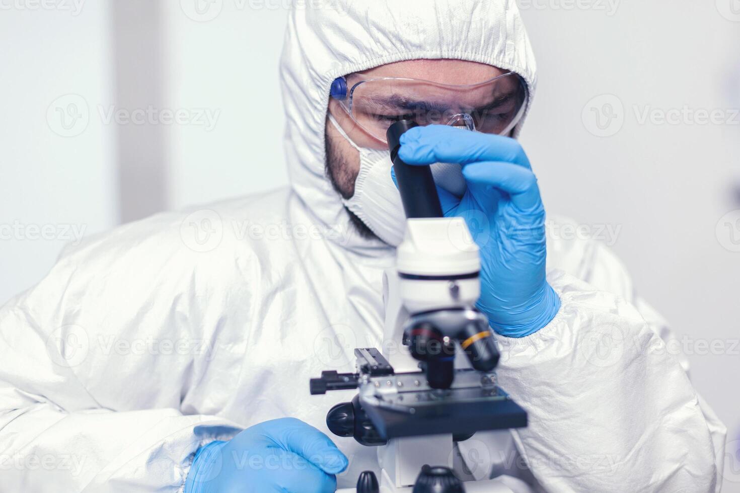 médico investigador mirando a través microscopio a virus muestra vestido en ppe traje. científico en protector traje sentado a lugar de trabajo utilizando moderno médico tecnología durante global epidemia. foto