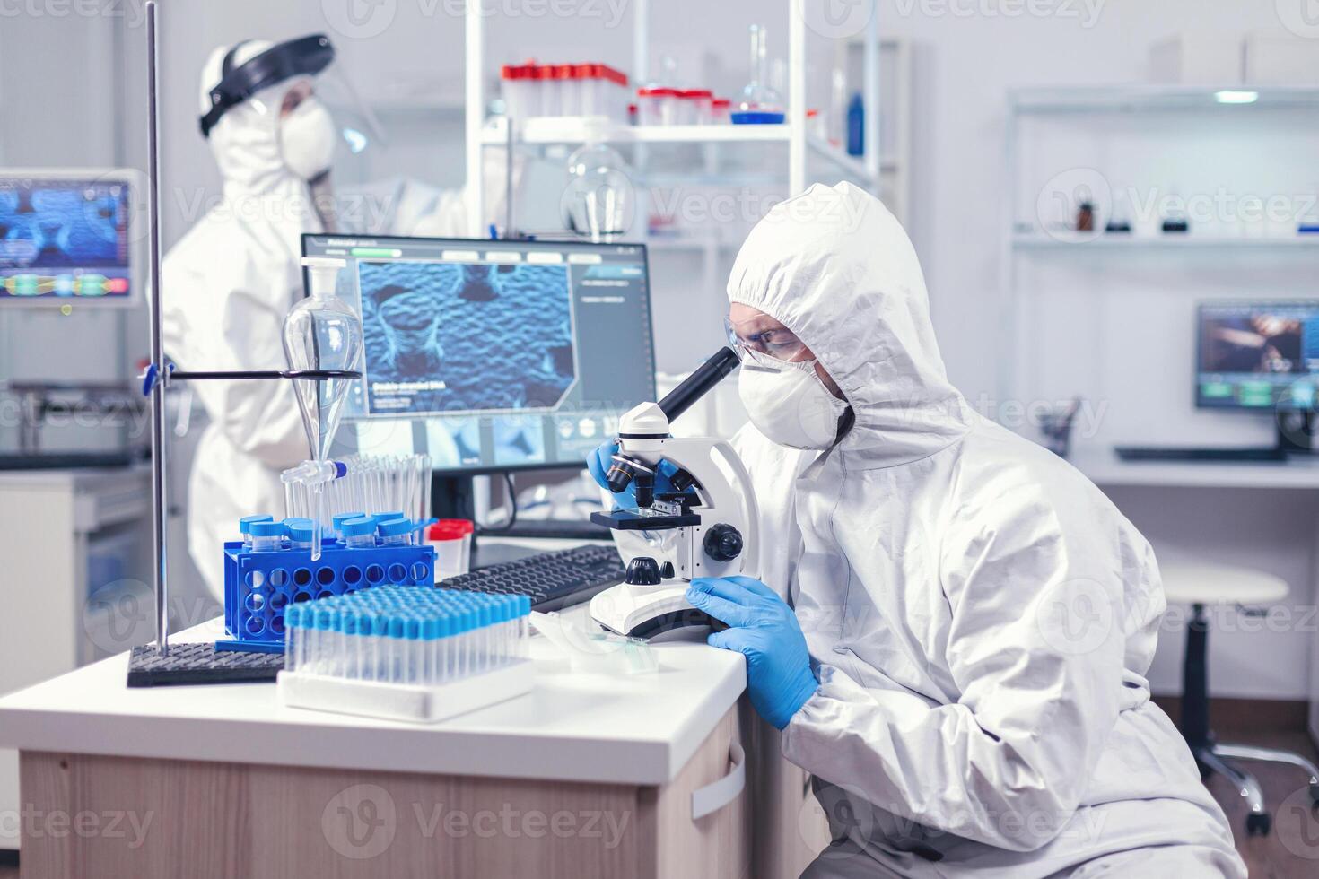 Man in ppe equipment conducting coronavirus investigation looking through investigation. Scientist in protective suit sitting at workplace using modern medical technology during global epidemic. photo