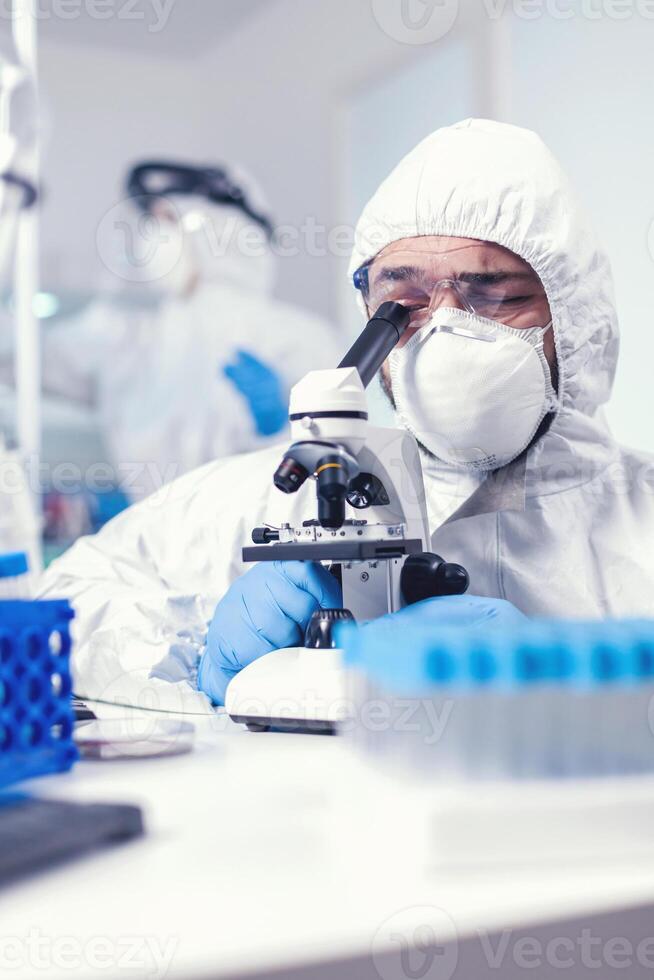 Medical researcher wearing coverall searching for covid treatment looking through microscope. Scientist in protective suit sitting at workplace using modern medical technology during global epidemic. photo