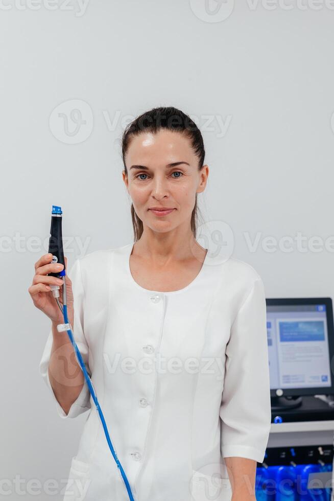 Middle aged beautician holding ultrasound device for face lifting and skin tightening procedure. Young woman cosmetologist using modern cosmetology equipment in clinic photo