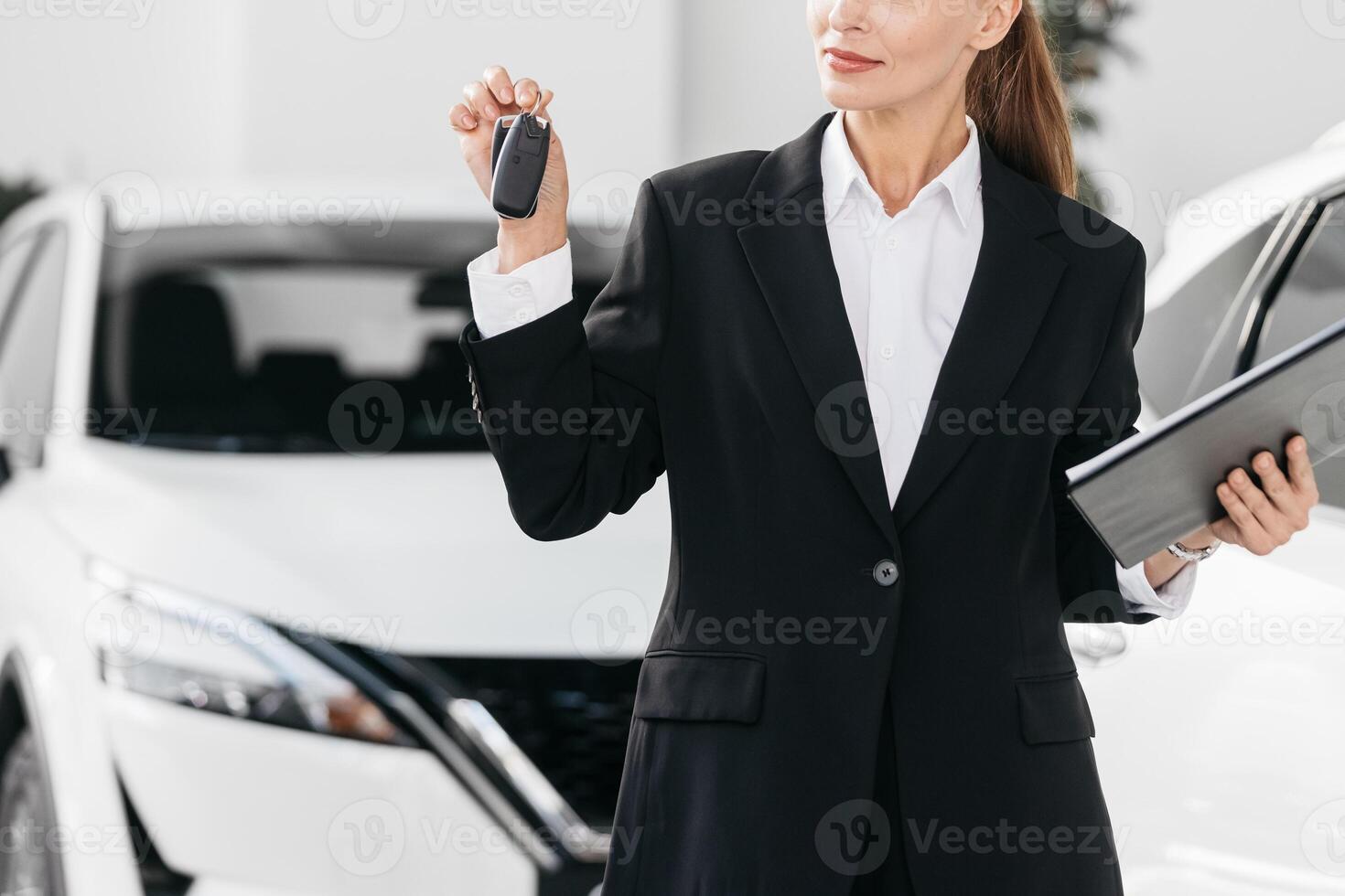 Female Competent car dealer in a suit with tablet at dealership center. Manager in auto dealership presents new cars. Dealer giving key to new owner in auto show or salon. photo