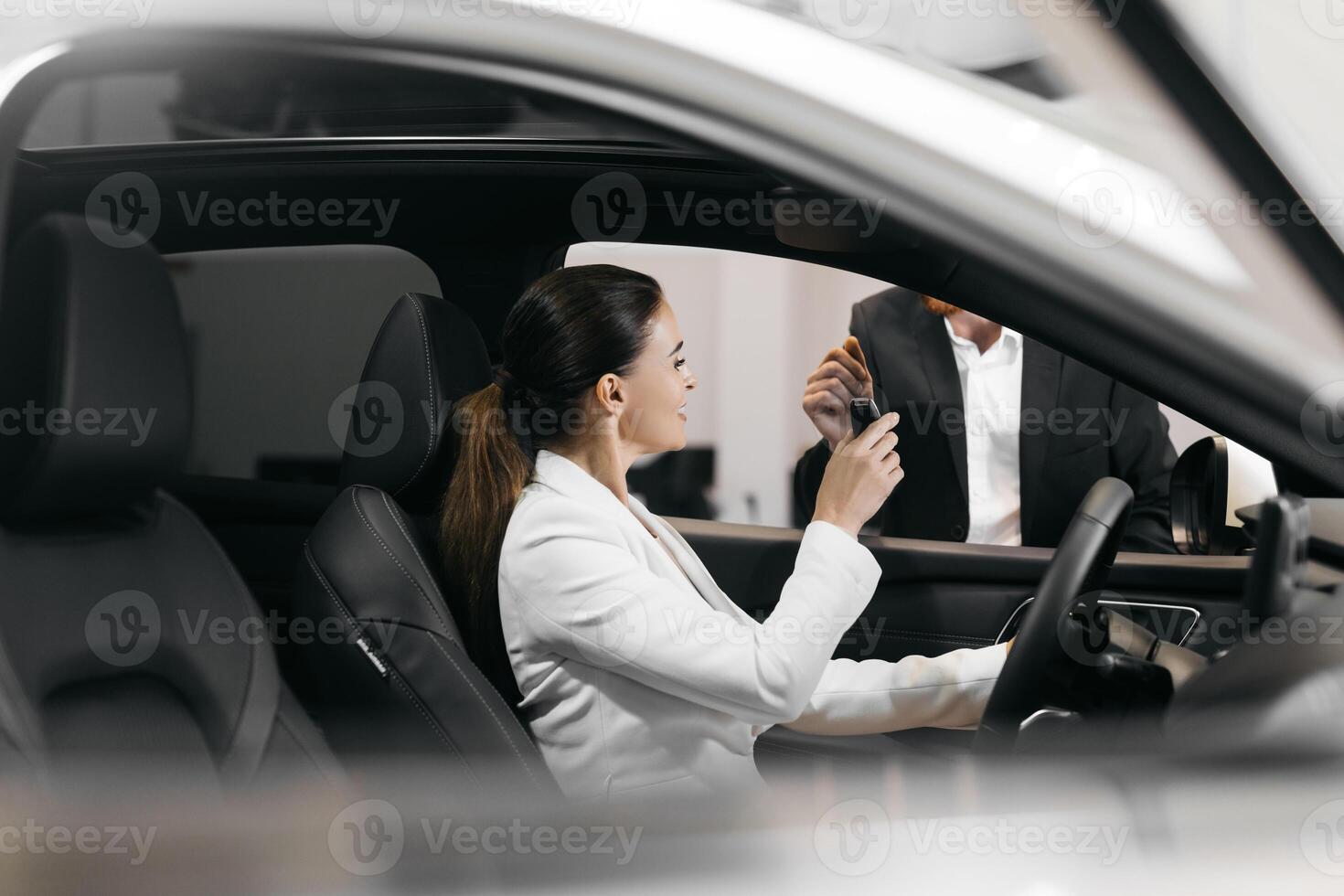 Car dealer giving keys to a young woman sitting inside the car. Joyful executive woman getting keys from new car. Happy auto owner buying transport at modern salon. photo