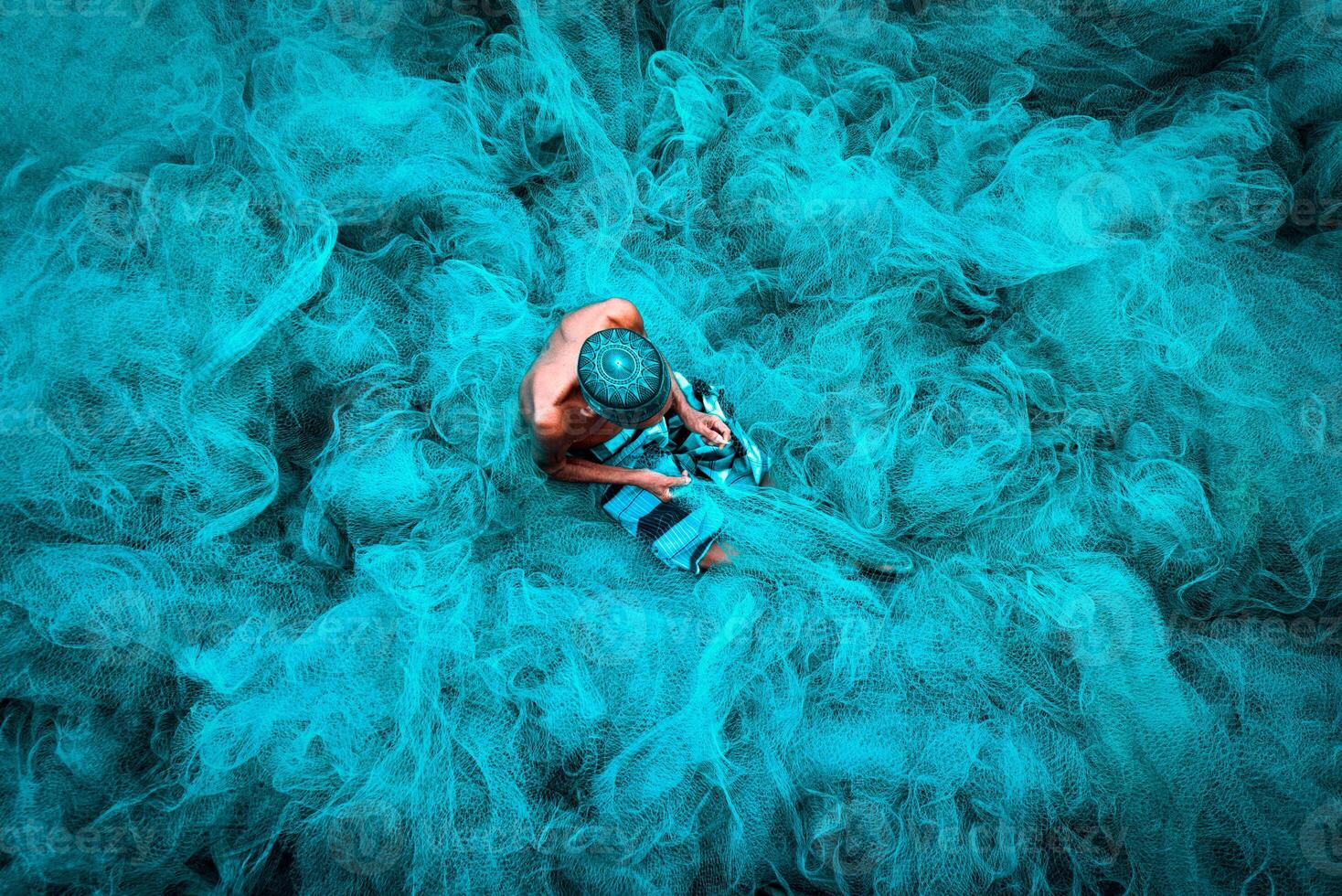 Old fisherman hands sewing blue fishing nets sitting on the ground and surrounded big net photo