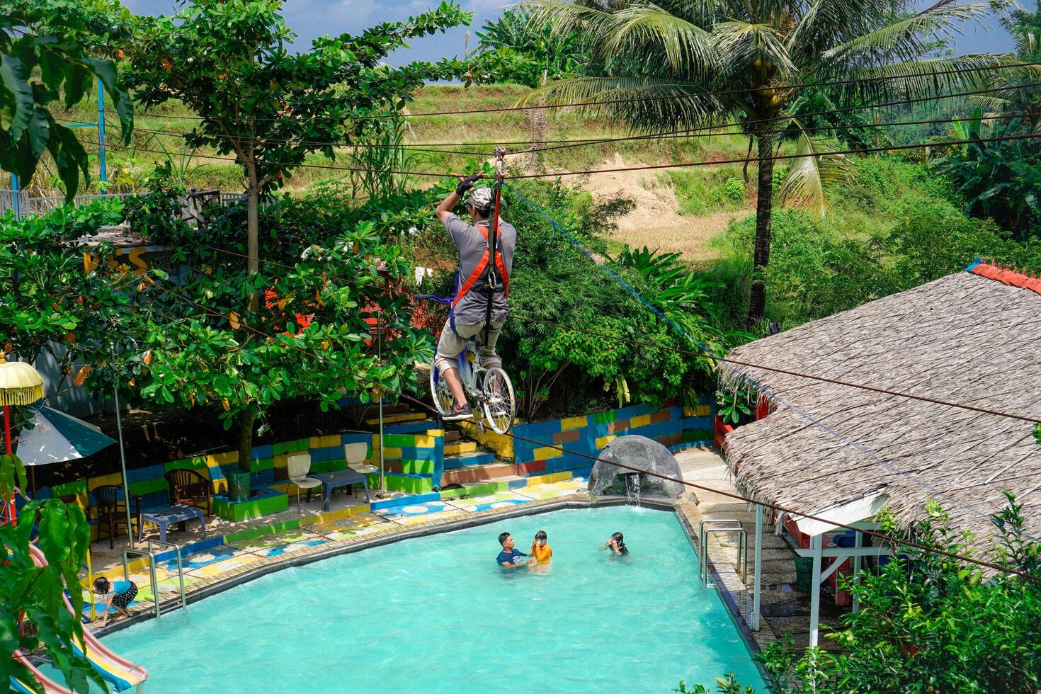 jepra jawa tengah, april 16, 2024 - rear view of a man on a bicycle designed to be a flying fox ride over a swimming pool surrounded by hills. photo