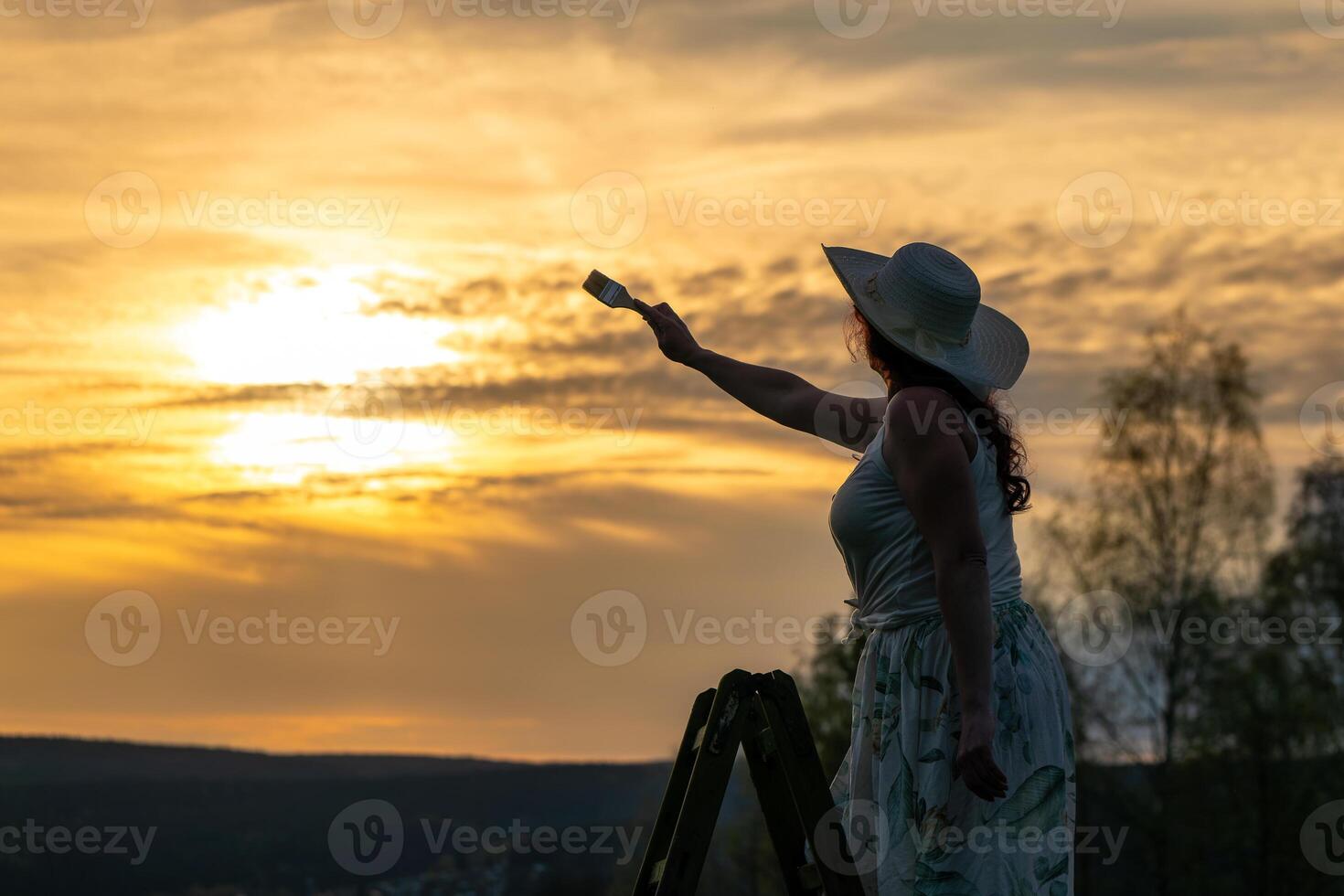 a woman in a dress stands on a wooden ladder and paints the sunset photo