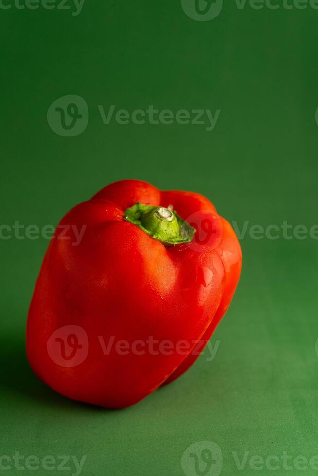 A shiny red pepper sits against a green backdrop, creating a vivid color contrast. The image highlights the pepper's smooth texture and rich hue, suggesting freshness and vibrancy. photo