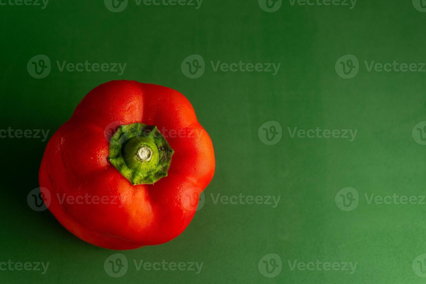 A shiny red pepper sits against a green backdrop, creating a vivid color contrast. The image highlights the pepper's smooth texture and rich hue, suggesting freshness and vibrancy. photo