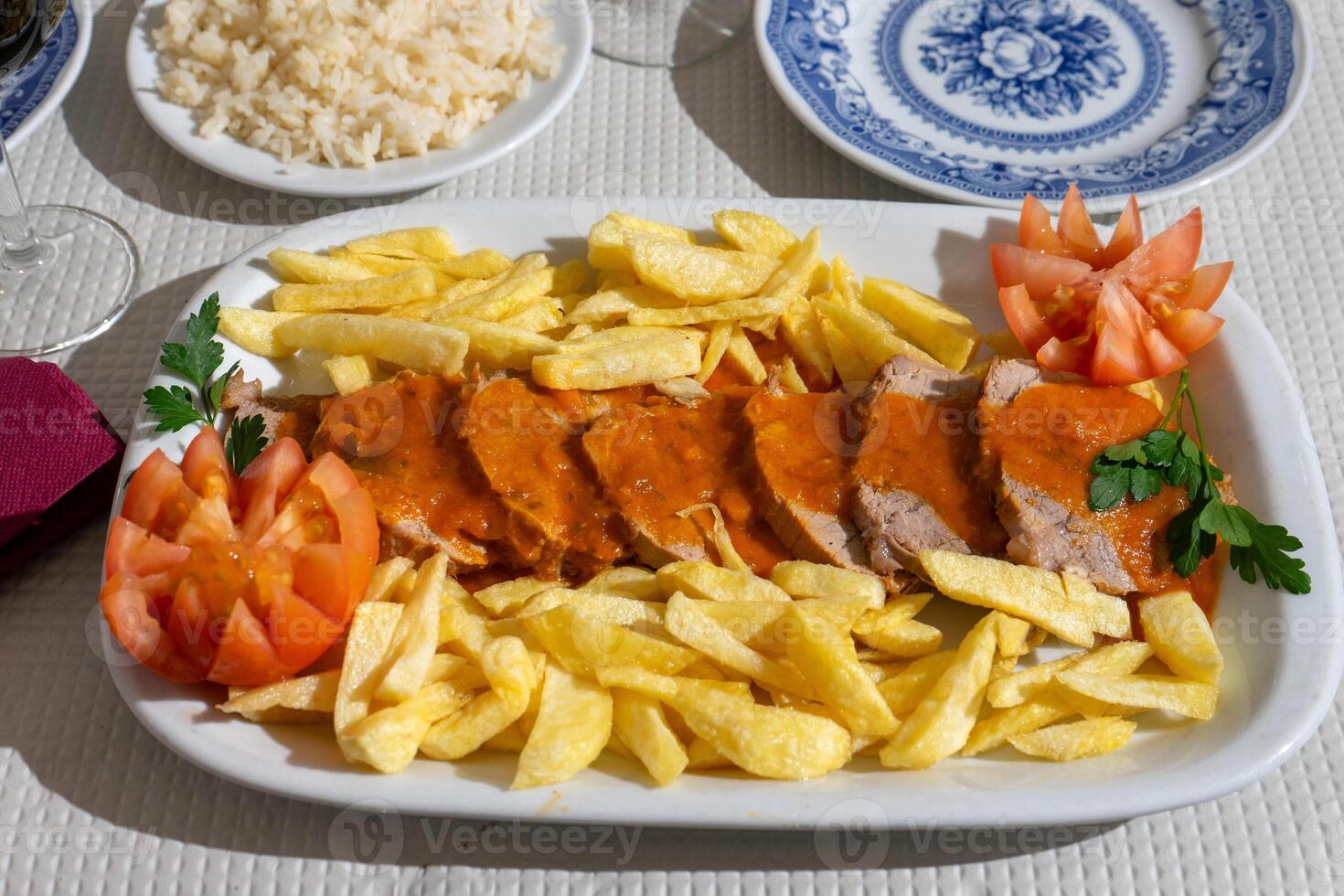 A plate of veal stew in a rich tomato sauce, served with crispy fries, is presented on a table at a traditional Alentejo restaurant. The rustic setting and hearty dish create a inviting atmosphere. photo