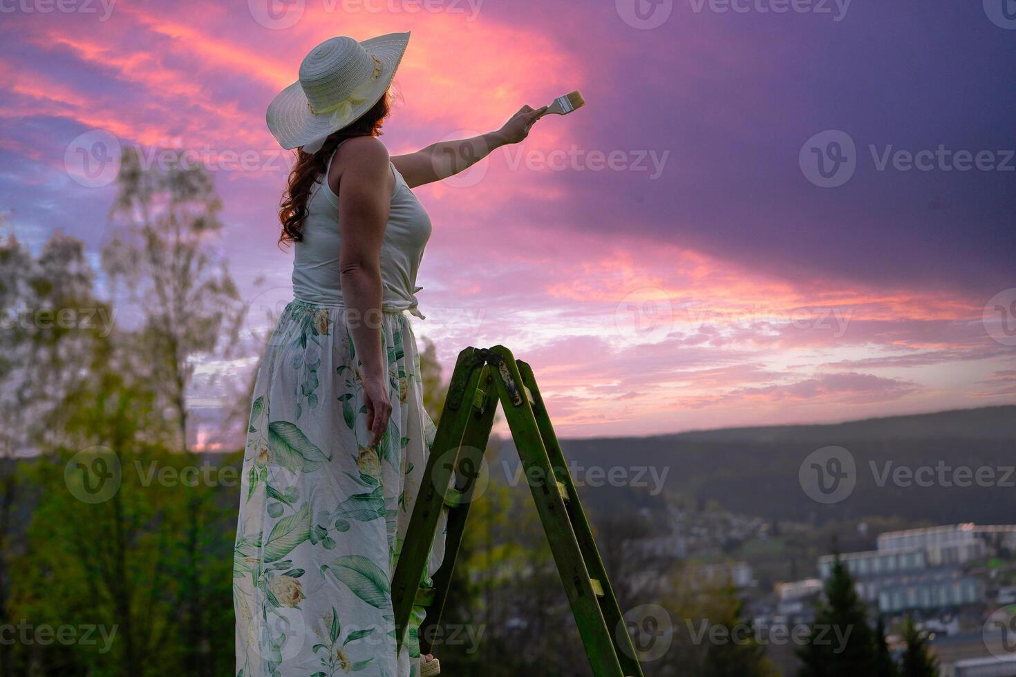 a woman in a dress stands on a wooden ladder and paints the sunset photo