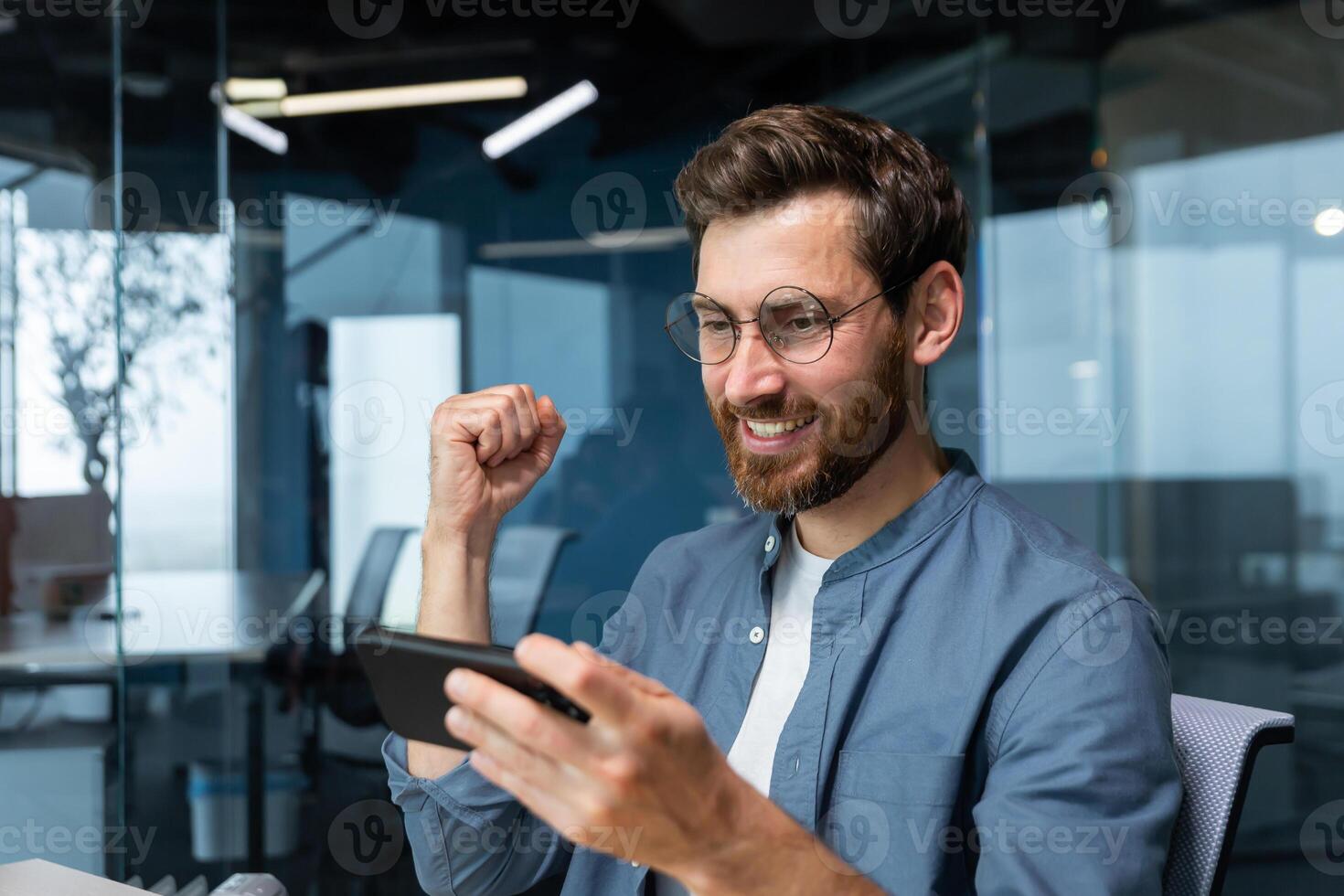 hombre dentro oficina a lugar de trabajo jugando juegos en teléfono inteligente, empresario contento con victorioso se regocija y sostiene mano arriba en triunfo gesto, jefe en camisa contento con logro y resultado. foto