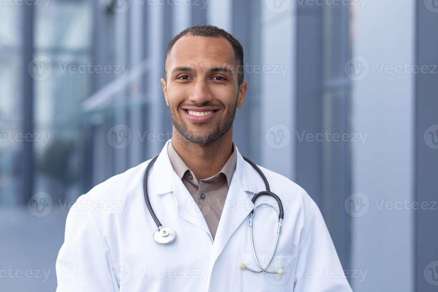 retrato de exitoso y sonriente africano americano doctor, hombre en médico Saco con estetoscopio mirando a cámara y sonriente, fuera de moderno hospital foto