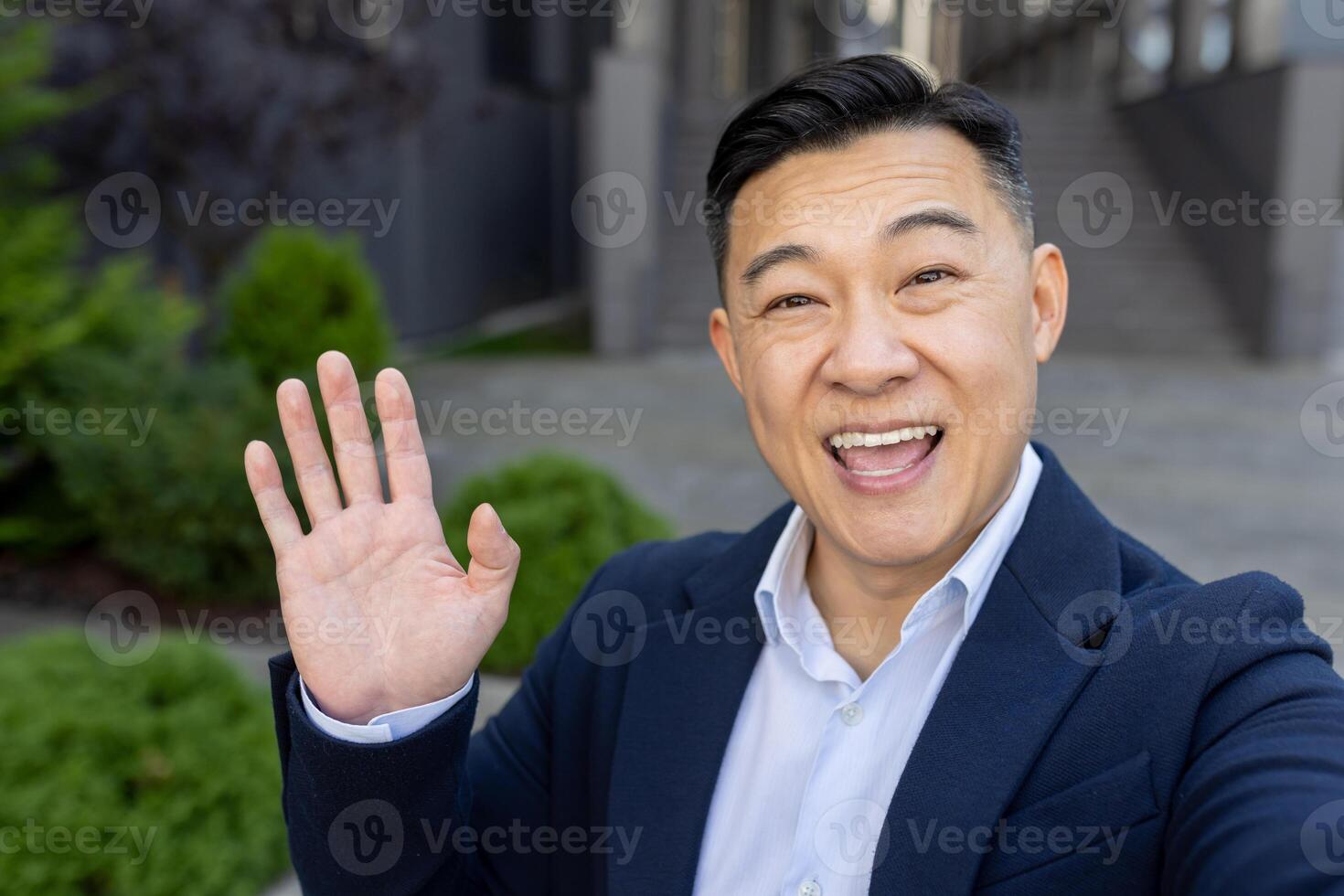 Close-up photo of a young Asian male businessman standing outside a building in a suit, talking on a call on the phone, greeting and waving at the camera.