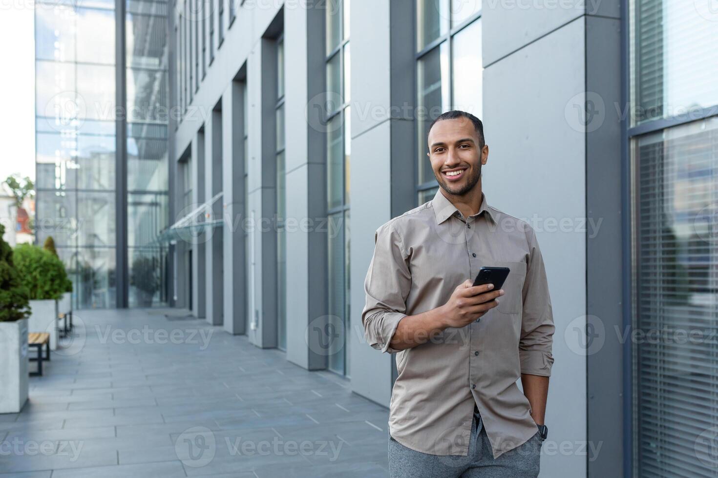 Hispano empresario caminando fuera de oficina edificio, hombre participación teléfono inteligente, sonriente y mirando a cámara foto