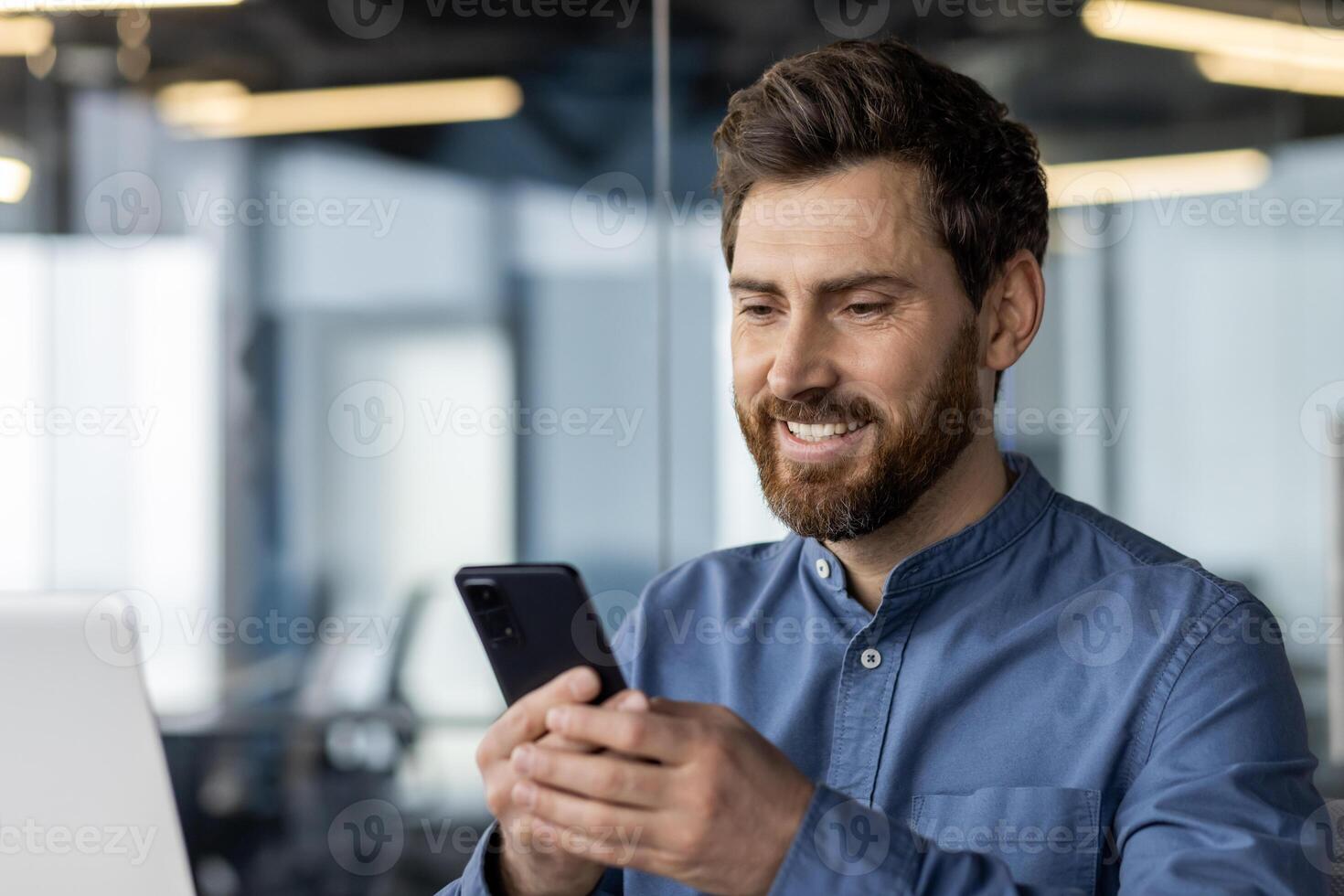 Close-up photo of a tired young man sitting at his desk in the office and using a mobile phone.