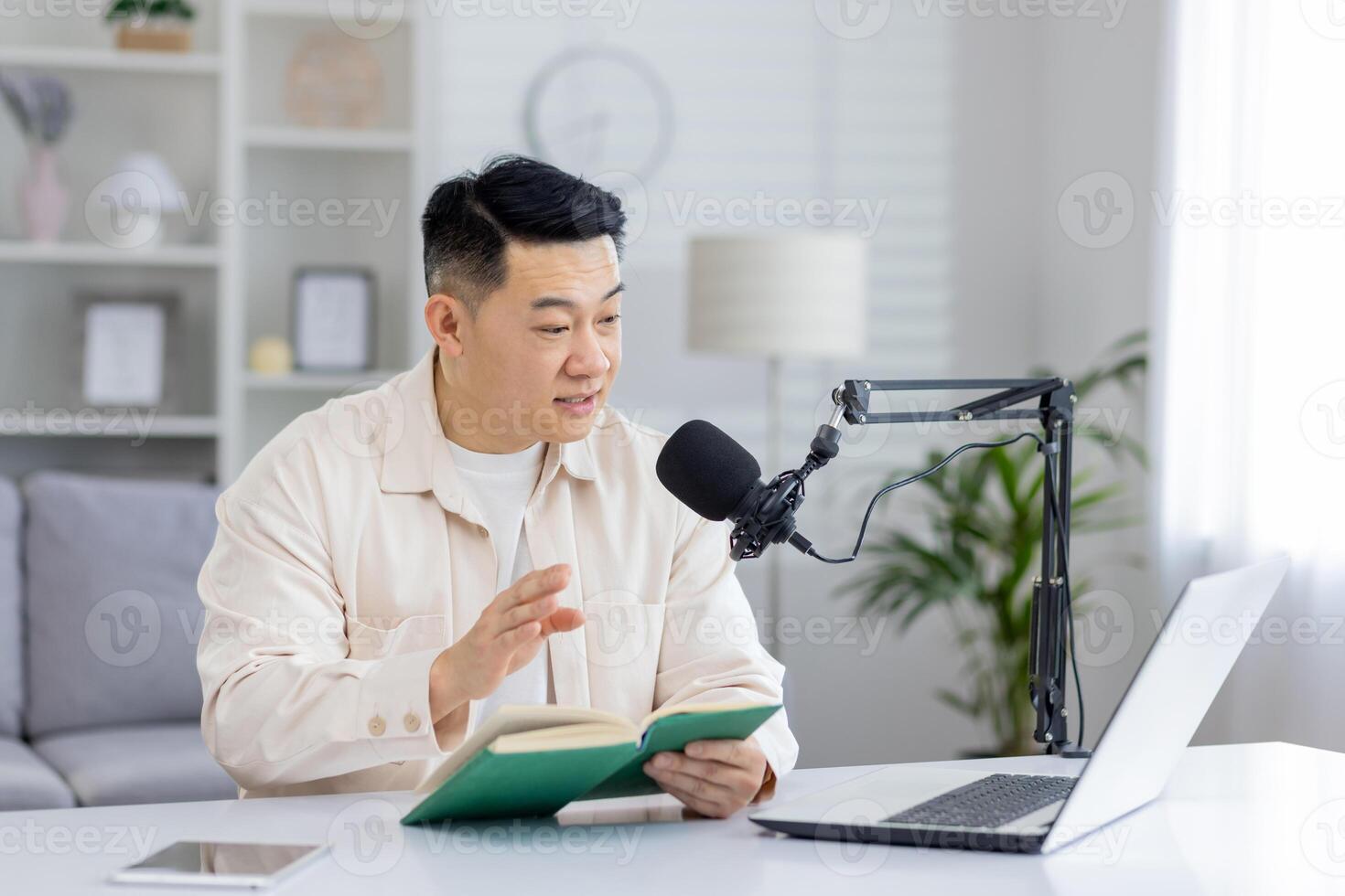 Engaged Asian man recording a podcast at his home studio, with a book open in hand and microphone setup on the desk. photo