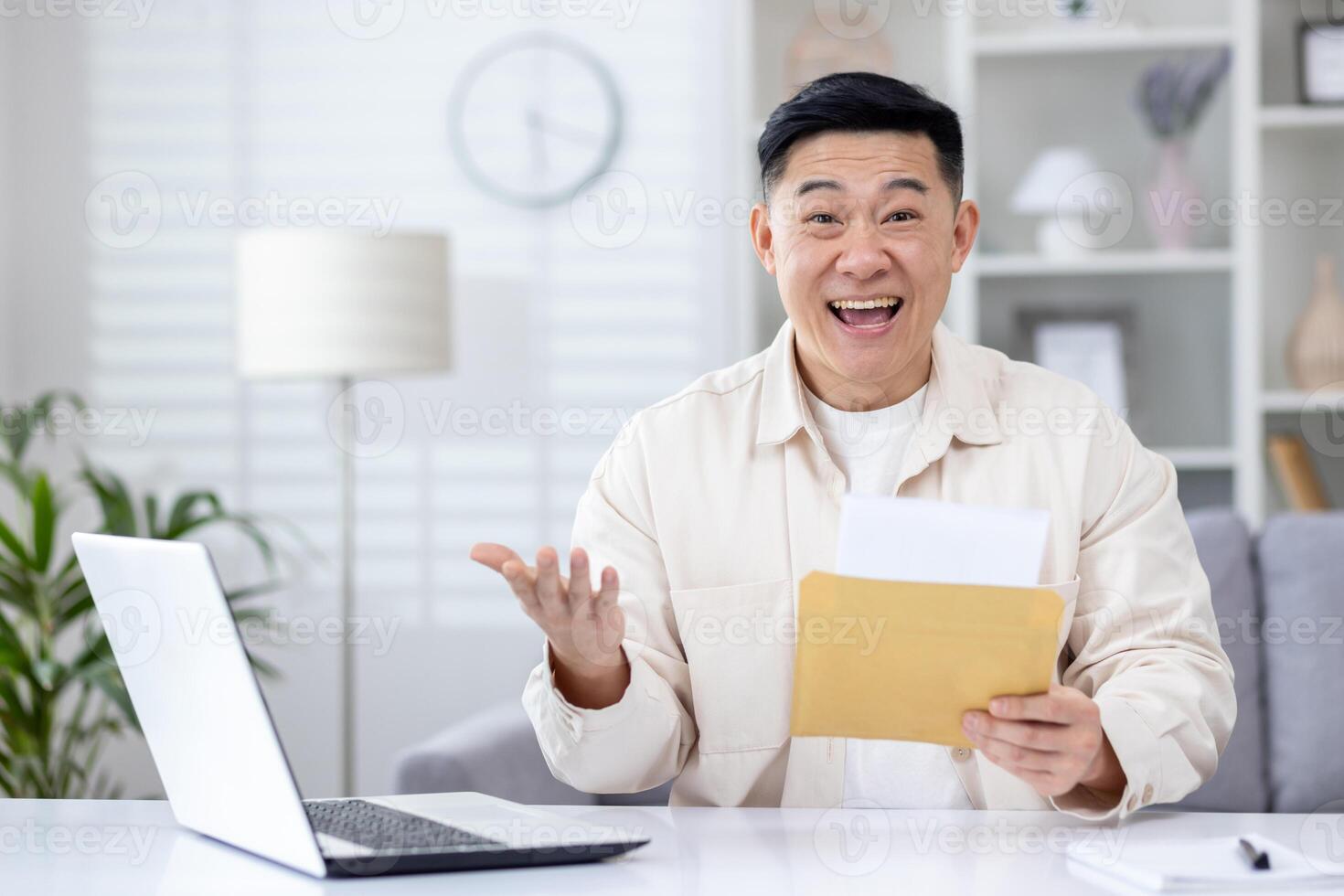 Surprised Asian man sitting at desk in home office, holding open envelope in hands, can't believe his luck. Korean senior man is happy with the win, good news, look at the camera. photo