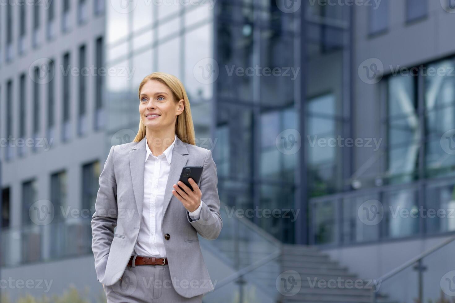 Inspired blonde lady with mobile phone wearing grey suit and looking away while keeping hand in pocket. Motivated businessperson going out for lunch break and checking route to nearest restaurant. photo