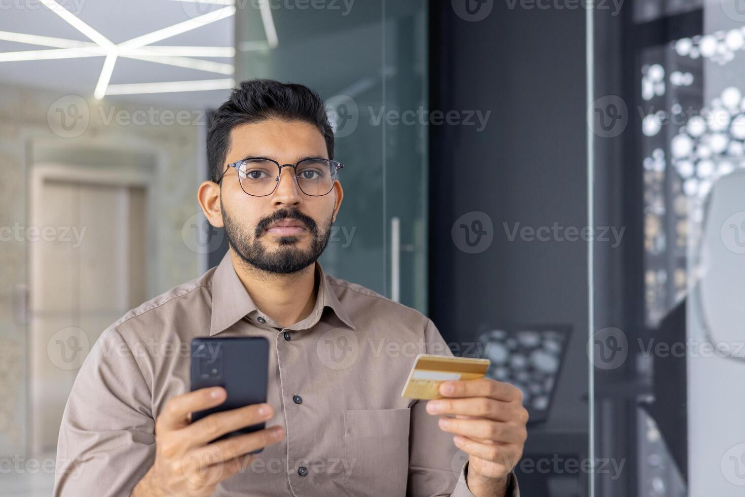 un enfocado hombre en lentes sostiene un crédito tarjeta y teléfono inteligente en un moderno interior configuración, posiblemente haciendo un en línea pago. foto