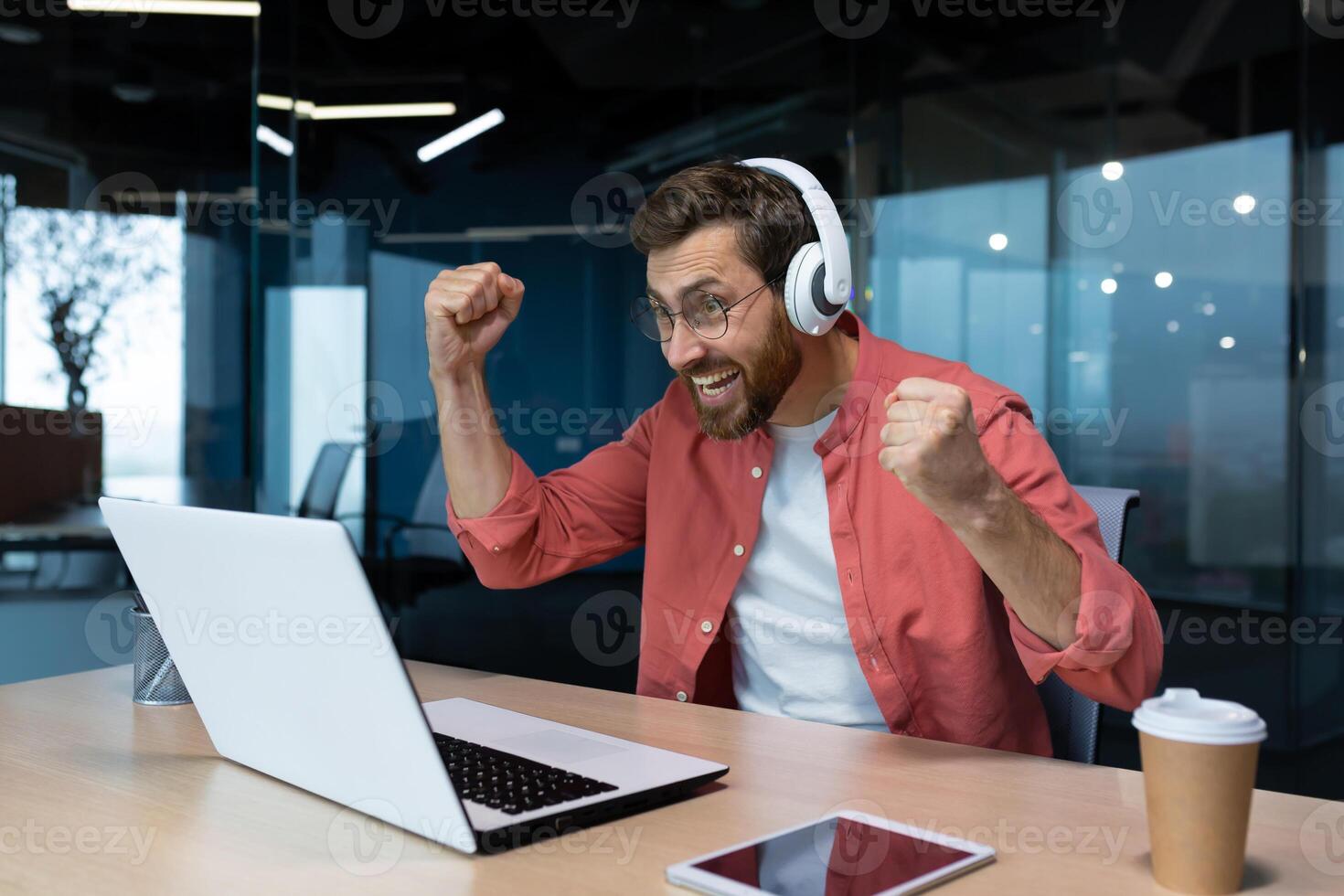 Man at the workplace inside the office is cheering watching a sports match on the computer, joyful in headphones, satisfied with the result of the sports team. photo