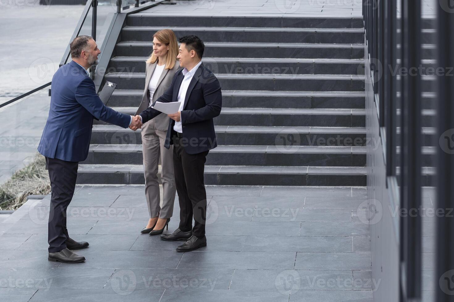 Meeting of three colleagues from outside the office building, experienced and mature IT specialists, greeting and shaking hands, business persons in business suits, diverse group of people photo
