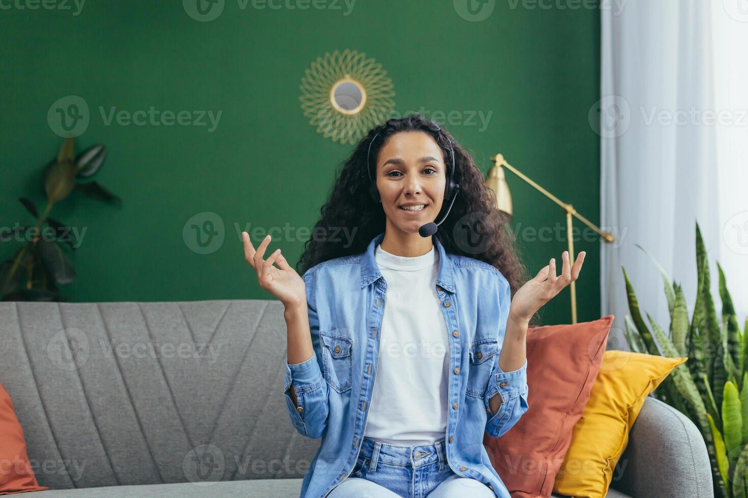 Joyful hispanic woman with call headset at home smiling and looking at camera, woman studying and working online remotely sitting on couch at home. photo