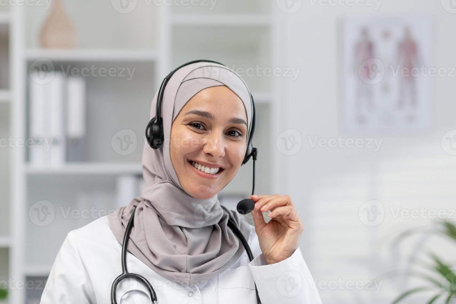 A cheerful Muslim female doctor in a hijab with a headset, ready for an online consultation in a bright medical office, looking at the camera. photo