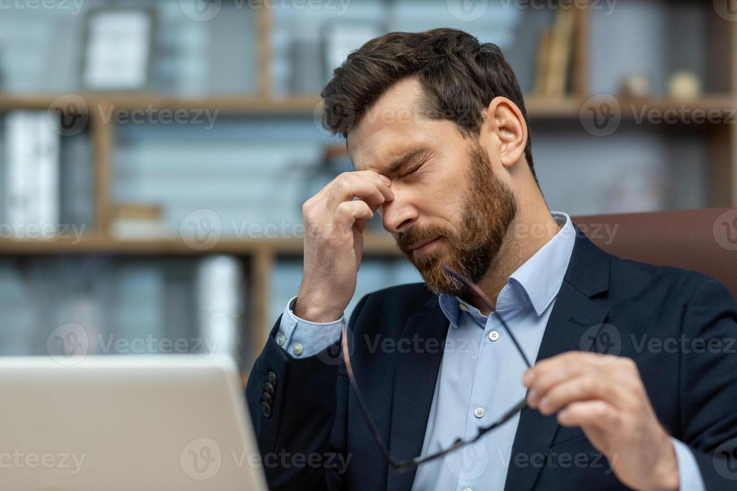 Serious mature man in business attire feeling fatigued, rubbing his eyes while focusing on his work in a classic home office setting. photo